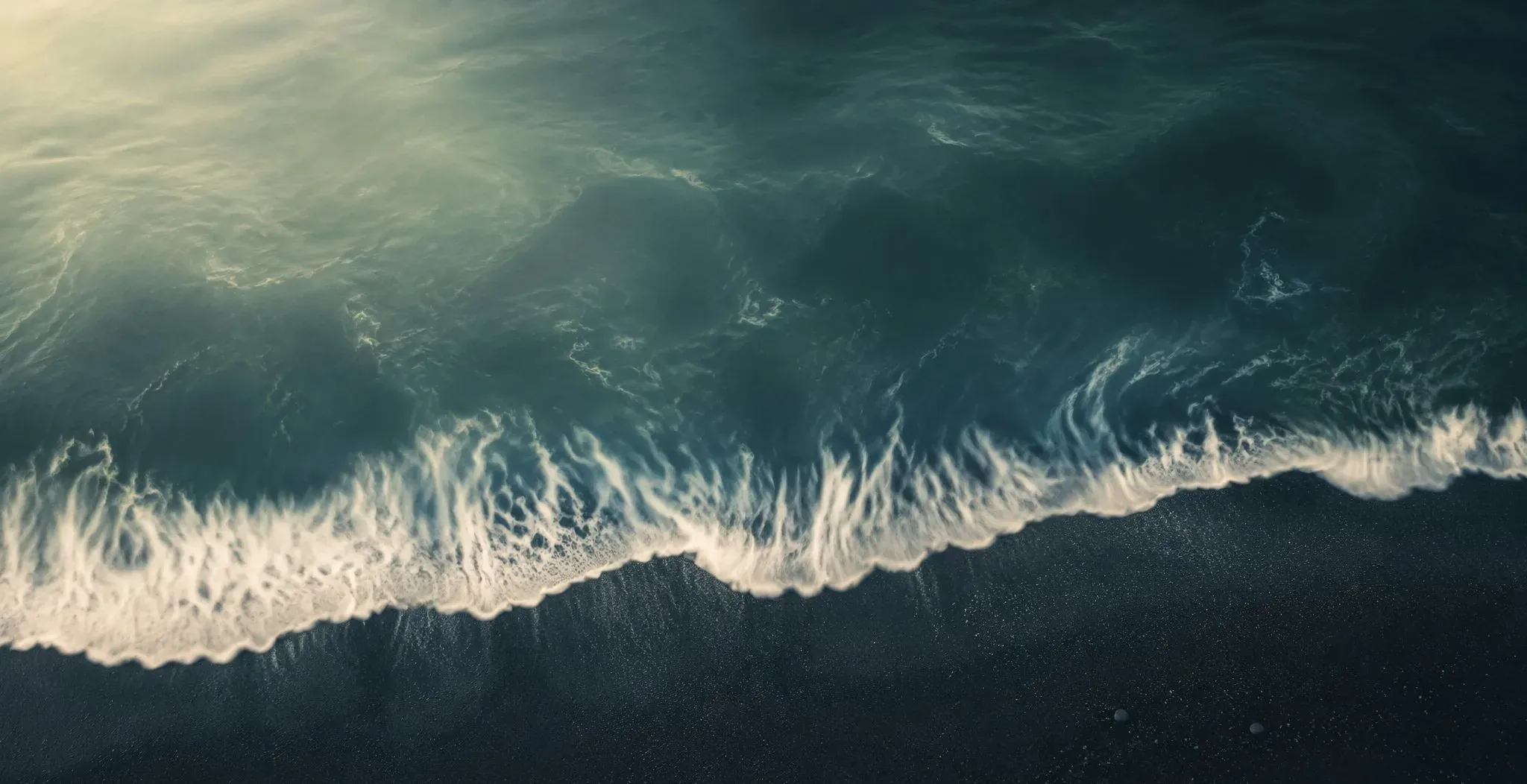 Aerial view of teal Pacific waves washing over dark volcanic sand